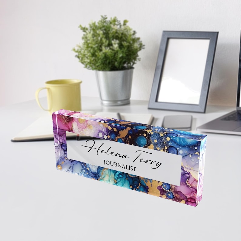 May include: A clear acrylic desk nameplate with an abstract design in shades of purple, blue, and gold. The nameplate reads "Helena Terry, Journalist".