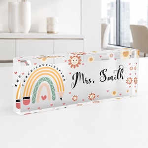 May include: A rectangular clear acrylic desk sign featuring the text Mrs. Smith in black script. The design includes a colorful pencil-shaped rainbow, small hearts, and smiling flower icons with the words teach, love, and inspire printed.
