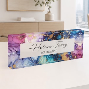 Könnte beinhalten: Ein rechteckiges Schreibtisch-Namensschild aus Acryl mit einem lebendigen Alkoholtintenmuster in Violett-, Blau- und Goldtönen. Ein weißes Querband in der Mitte zeigt den Namen Helena Terry in schwarzer Schrift und den Titel JOURNALIST.
