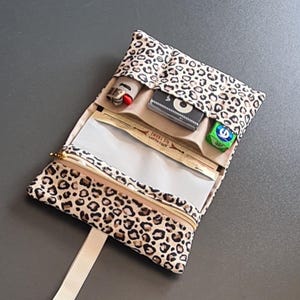 Può includere: Una custodia in tessuto arrotolabile con motivo leopardato nei toni del marrone, nero e crema. L'interno presenta diverse tasche contenenti un accendino, una piccola scatola rettangolare, un bastoncino sottile e un cubo verde. Cinturino elastico beige.