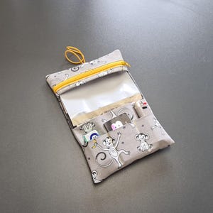 Può includere: Un organizer in tessuto grigio con cerniera gialla e tasca in plastica trasparente. L'organizer presenta un motivo ripetuto di scimmie bianche dei cartoni animati. Ha più tasche e un anello giallo.