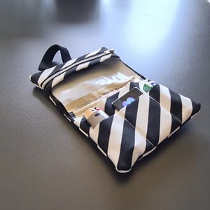 Könnte beinhalten: Eine Stofftasche mit schwarz-weißen Diagonalstreifen, ausgestattet mit einem Reißverschlussfach mit transparentem Kunststofffenster, einer schwarzen Trageschlaufe und mehreren Innentaschen, die ein Feuerzeug und kleine Gegenstände enthalten.