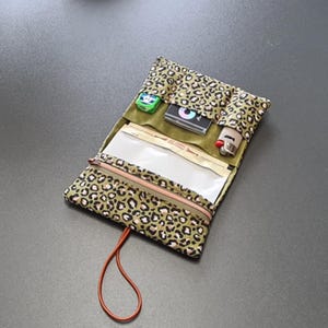Puede incluir: Un estuche de tela con estampado de leopardo que presenta una parte superior abierta con tres bolsillos interiores que contienen una caja verde pequeña, un objeto rectangular negro y un encendedor. Incluye un compartimento con cremallera y un cordón naranja.
