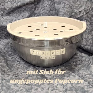 May include: A stainless steel bowl with a beige lid. The bowl has a label that reads "Popcorn Only". The lid has holes for steaming popcorn. The bowl is on a grey background with the text "mit Sieb für ungepopptes Popcorn".