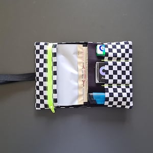 Puede incluir: Un estuche de tela con estampado de cuadros blancos y negros, cremallera de color amarillo neón, una cinta elástica negra y un compartimento interior con tres bolsillos verticales que contienen una caja verde, una tarjeta negra y un encendedor azul.