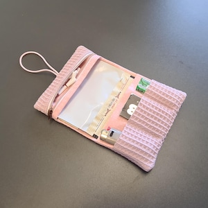 Könnte beinhalten: Eine rosa Tasche mit Waffelstruktur, Reißverschluss und Schlaufe. Die Tasche ist geöffnet und zeigt eine transparente Plastiktasche, ein Feuerzeug und einige kleine Gegenstände. Der Text "FRESH LOCK SMART ZIP" ist sichtbar.