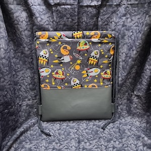 May include: A gray drawstring bag with a gray and yellow fabric lining featuring a space-themed pattern with rockets, dinosaurs, and stars. The bag has a gray leather bottom.