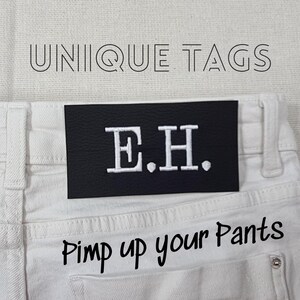 Peut inclure: Un jean en denim blanc avec un patch en cuir noir qui dit "E.H." et le texte "Pimp up your pants" imprimé sous le patch.