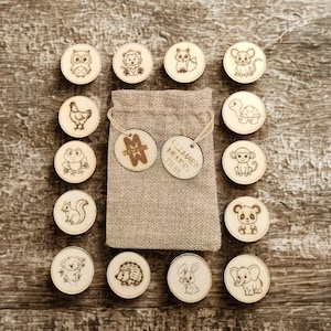 Puede incluir: Un juego de memoria de madera con ilustraciones de animales grabadas, como un búho, un león y un zorro. El juego incluye una bolsa de tela y dos etiquetas de madera con texto. Las piezas del juego están dispuestas sobre una superficie de madera.