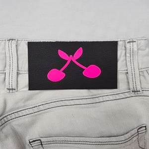 May include: A black leather patch with two pink cherries on a pair of light gray jeans.