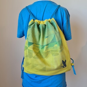 Könnte beinhalten: Ein gelb-grüner Kordelzug-Rucksack mit geometrischem Design. Die Tasche hat blaue Kordelzüge und eine kleine schwarze Grafik eines Surfbretts und Wellen. Der Rucksack wird über einem blauen T-Shirt getragen.