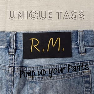Può includere: Una toppa in pelle nera con le iniziali "R.M." ricamate in oro cucite su un paio di jeans blu. Il testo "Pimp up your Pants" è ricamato sulla tasca posteriore dei jeans.