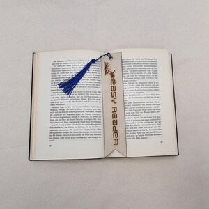 Könnte beinhalten: Ein Holz-Lesezeichen mit einem blauen Quasten und dem Text "Easy Reader" in Weiß. Das Lesezeichen hat ein dekoratives Design mit einem Schmetterling und Blumen.