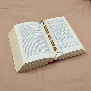 Könnte beinhalten: Ein offenes Buch mit einem Holzlesezeichen. Auf dem Lesezeichen steht in einer kräftigen Schriftart "READ OR DIE". Am unteren Ende des Lesezeichens befindet sich ein kleines dekoratives Design. Die Seiten des Buches sind mit Text gefüllt.