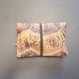 May include: A rectangular zippered pouch featuring a brown and tan tortoise shell pattern across the entire surface. A thin, dark olive green elastic band is wrapped vertically around the center of the pouch, and a small gold zipper pull is visible.
