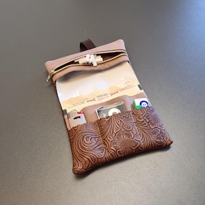 Op de afbeelding: Een bruine leren etui met een reliëf bloemenpatroon op het voorvak. De etui bevat een bovenste vak met ritssluiting met witte voorwerpen, een doorzichtig plastic gedeelte en een open vak met een aansteker en kleine voorwerpen.