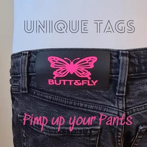 Könnte beinhalten: Schwarze Jeans mit einem schwarzen Lederaufnäher mit einem pinkfarbenen Schmetterlingsdesign und dem Text "BUTT&FLY". Das Bild enthält auch den Text "UNIQUE TAGS" und "Pimp up your Pants."