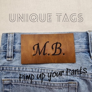 Könnte beinhalten: Nahaufnahme einer hellblauen Jeans mit einem braunen Lederetikett. Das Etikett ist mit den Initialen "M.B." in schwarzer Schrift geprägt. Der Text "UNIQUE TAGS" und "Pimp up your Pants" sind ebenfalls sichtbar.