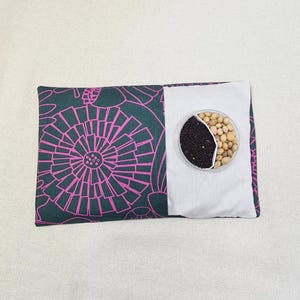 May include: A dark green and pink patterned fabric pouch with a white fabric pocket. The pocket contains a small white container with two compartments, one filled with black seeds and the other with beige seeds.