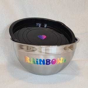 Könnte beinhalten: Eine Edelstahl-Schüssel mit einem schwarzen Deckel. Die Schüssel hat einen Regenbogen-Aufkleber mit der Aufschrift "RAINBOWL". Auf dem Deckel befindet sich ein kleiner lila-blauer Herzaufkleber.