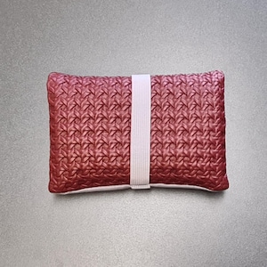 Pode incluir: Uma bolsa retangular com um exterior vermelho profundo e um padrão texturizado de estilo entrelaçado. Uma faixa elástica vertical de cor clara está posicionada no centro da bolsa, mantendo os dois lados unidos num design simples e funcional.