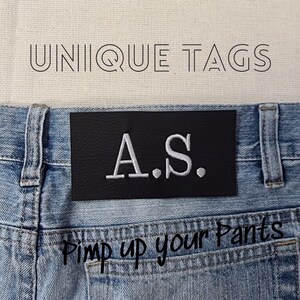 May include: A black leather patch with white stitching that says "A.S." is sewn onto a pair of blue denim jeans. The text "Pimp up your Pants" is written in black on the jeans below the patch.