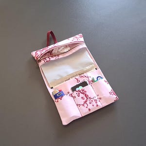 以下が含まれることがあります： 淡いピンク色の布製オーガナイザーポーチで、濃いピンク色の桜の模様が施されています。ポーチには上部のファスナー付きコンパートメント、透明なプラスチック製のセクション、ライターや小物を収納できる3つのオープンポケットが付いています。