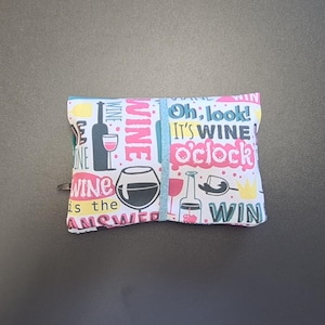 May include: A rectangular fabric pouch featuring a white background with a colorful pattern of wine bottles, glasses, and text including "Oh, look! It's WINE o'clock" and "WINE is the ANSWER" in pink, yellow, teal, and black ink with a blue elastic band.