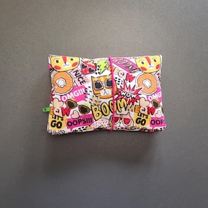 Puede incluir: Una bolsa rectangular de tela con un estampado vibrante de estilo caricatura. El diseño presenta caras de gato, rosquillas, gafas de sol y bocadillos con frases como "BOOM" y "OMG!!!". Una banda elástica rosa asegura la bolsa, y un clip verde está adjunto.