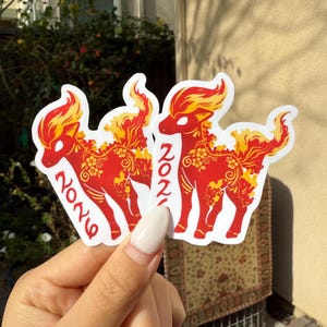 May include: Red and yellow stickers with a deer design, floral details, and the number "2021". The stickers have a white border and are held in a hand.