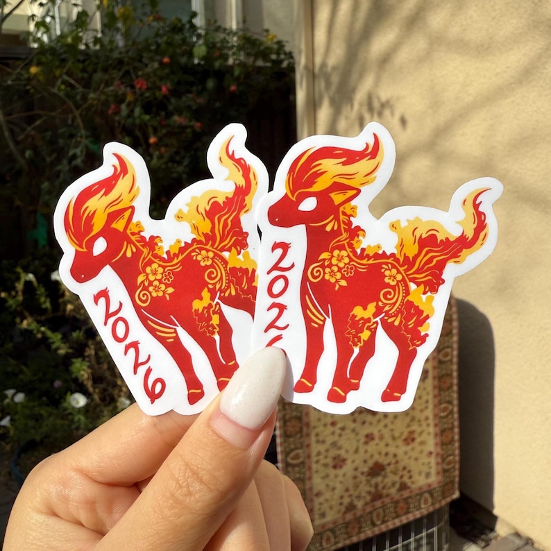 May include: Red and yellow stickers with a deer design, floral details, and the number "2021". The stickers have a white border and are held in a hand.