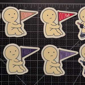 May include: A set of eight die-cut stickers featuring cartoon figures in various poses. Some figures hold pennants labeled Senators, Ottawa, Frost, or Minnesota, while others wear jerseys or hold hockey sticks with pucks on a black grid cutting mat.