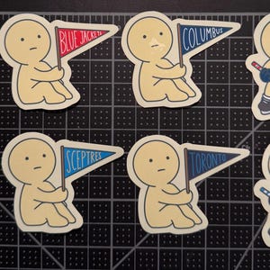 May include: A set of eight die-cut stickers featuring cartoon figures in various poses. Some figures hold pennants labeled Blue Jackets, Columbus, Sceptres, or Toronto, while others wear jerseys or hold hockey sticks with pucks on a black grid mat.