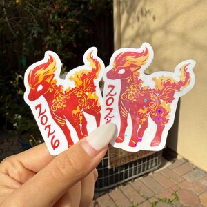 May include: Two stickers featuring a red mythical creature with yellow and orange accents. The stickers have the number "2026" printed on them. The stickers are held in front of a blurred green background.