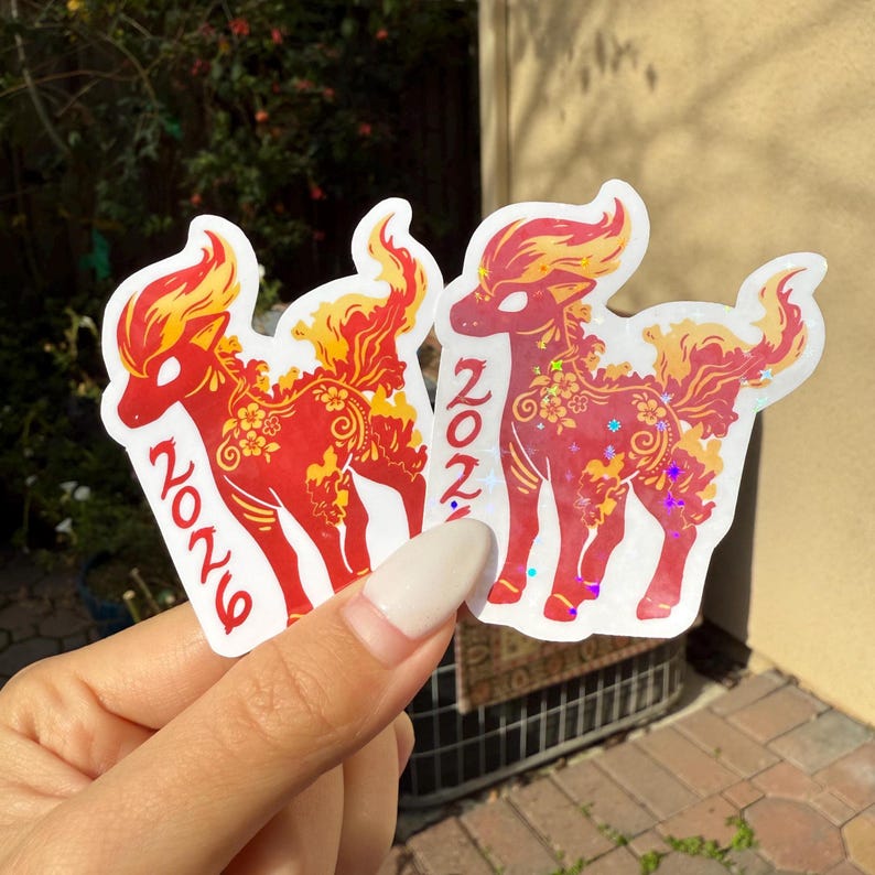 May include: Two stickers featuring a red mythical creature with yellow and orange accents. The stickers have the number "2026" printed on them. The stickers are held in front of a blurred green background.