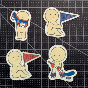 May include: Four die-cut stickers featuring a pale yellow cartoon character. One pulls on a blue and orange hockey jersey, one holds a blue pennant with the text ISLANDERS, one holds a red pennant with the text NEW YORK, and one holds a hockey stick.