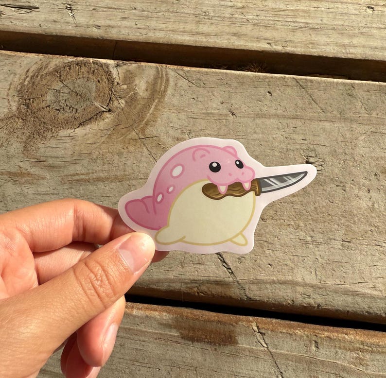 Puede incluir: Una pegatina rosa y crema con un dibujo de una foca sosteniendo un cuchillo en la boca. La foca tiene ojos oscuros y manchas en la cabeza y la espalda. El cuchillo es plateado con un mango negro.