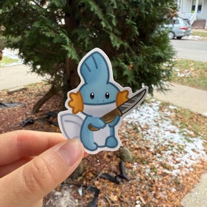 Op de afbeelding: Een blauw en wit cartoonfiguur met oranje vinnen die een zilveren mes vasthoudt. Het figuur is een sticker.