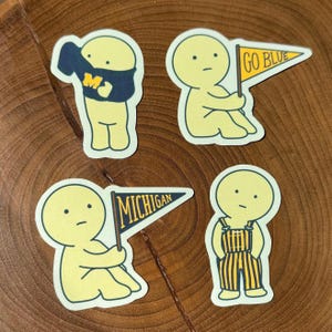 Op de afbeelding: Vier gestanste stickers met gele cartoonfiguren. Eén figuur draagt een marineblauw sweatshirt met een gele "M". Een ander houdt een gele wimpel met "GO BLUE". De derde houdt een wimpel met "MICHIGAN". De laatste figuur draagt een gele en zwarte overall.