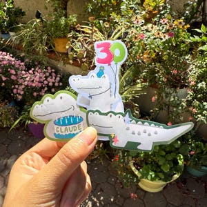 May include: Three alligator-shaped birthday decorations. One alligator holds a cake that says "CLAUDE". Another alligator holds a "30" balloon. The third alligator has a star. The decorations are white with green and blue accents.