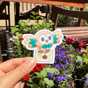 May include: A sticker featuring a cartoon owl with orange, teal, and cream colors. The owl is holding a cream-colored envelope with a brown wax seal. The sticker is held in front of a background of flowers and a wooden bench.