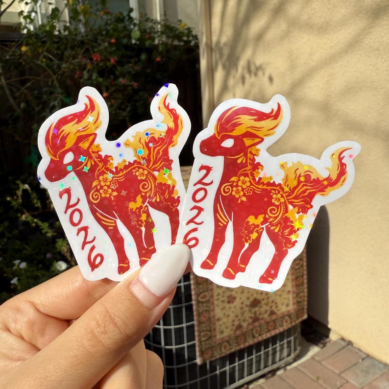 May include: Two red and gold stickers with a deer design and the number "2026". The deer has a fiery mane and tail, with floral and star accents. The stickers are held against a backdrop of foliage.