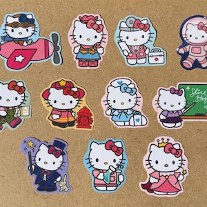 Peut inclure: Une collection d'autocollants découpés colorés représentant Hello Kitty dans diverses professions et costumes. Les motifs incluent un astronaute, un médecin, un pilote et un magicien. Les autocollants sont sur une surface brun clair.