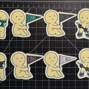 May include: A set of eight die-cut stickers featuring pale yellow cartoon figures. Four figures hold pennants labeled Stars, Dallas, Kings, and Los Angeles. Two figures wear hockey jerseys, and two figures hold hockey sticks with pucks.