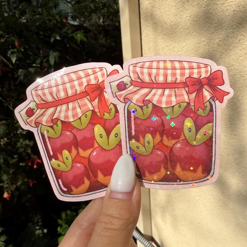 Op de afbeelding: Twee stickers met een pot appels. De potten hebben een rood-wit geruit deksel, een rode strik en zijn gevuld met rode appels met groene bladeren. De stickers hebben een roze rand.
