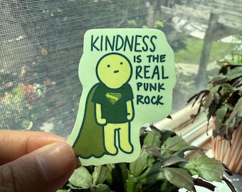 以下が含まれることがあります： 「Kindness is the Real Punk Rock」の文字と、マントを着た漫画のキャラクターが描かれたダイカットステッカー。キャラクターは黄色のエンブレムが付いた濃い緑色のシャツを着ています。ステッカーは明るい緑色の背景です。