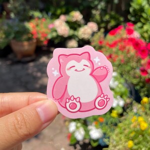May include: A pink and white sticker featuring a cartoon pink Pokemon character with a white belly and a happy expression. The character is sitting with its arms outstretched and has a white star on each side.