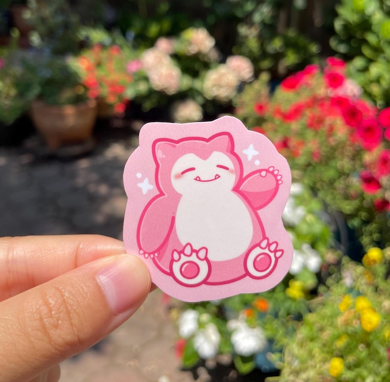 May include: A pink and white sticker featuring a cartoon pink Pokemon character with a white belly and a happy expression. The character is sitting with its arms outstretched and has a white star on each side.