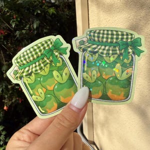 Op de afbeelding: Twee stickers met een potontwerp met groene en oranje vruchten. De potten hebben een groen-wit geruit deksel met een groene strik. De vruchten in de potten hebben een groen-oranje kleurenschema.
