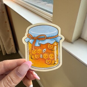 May include: A sticker featuring a jar of honey with a blue lid and a tied orange string. Inside the jar are cartoon honeycombs with smiling faces. The sticker is held up by a hand with a pink nail.